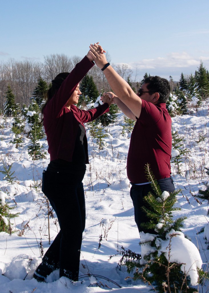 A couple dancing joyfully in a snowy field surrounded by evergreen trees, holding hands and smiling at each other.