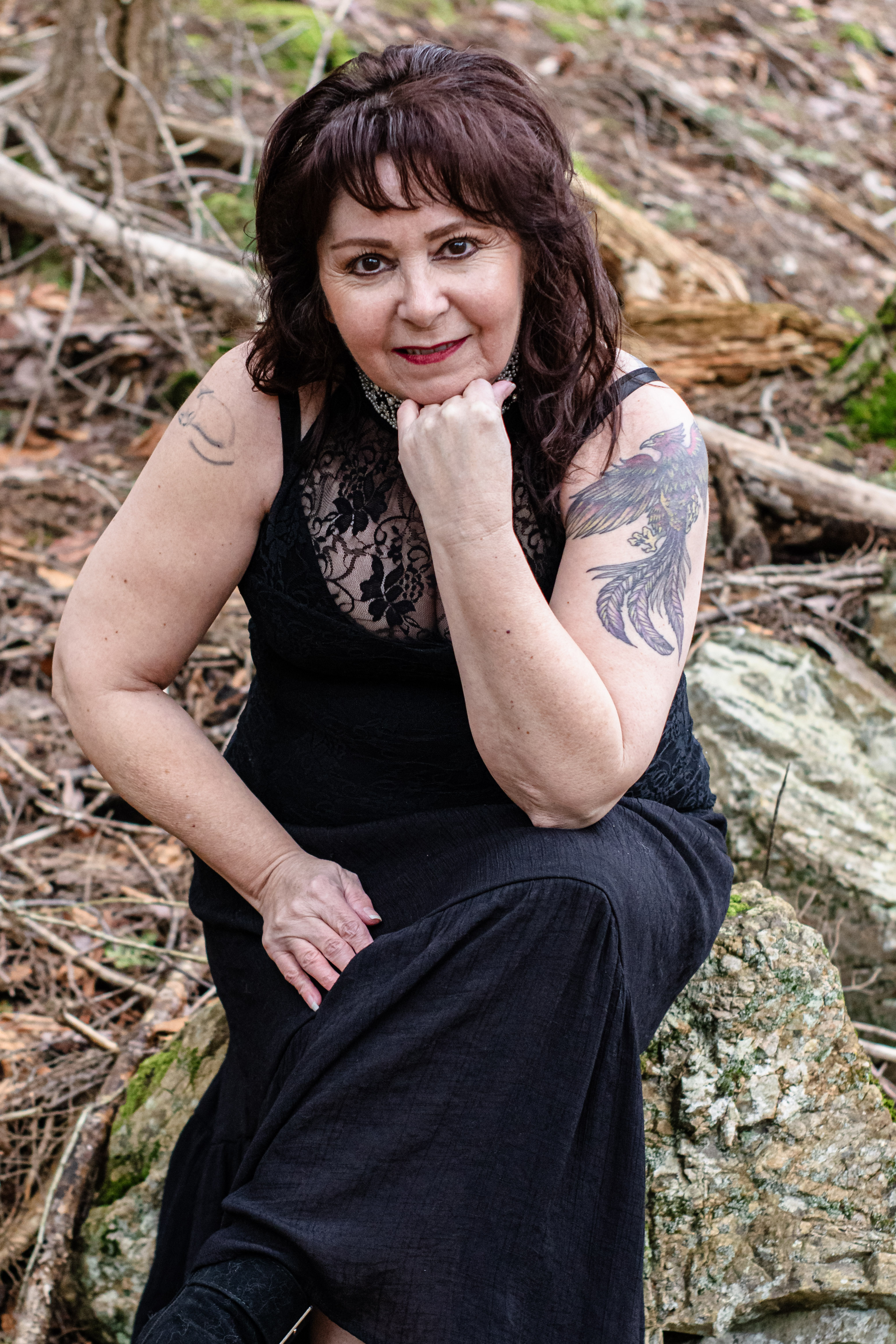 A woman sitting on a rock in a forest, wearing a black lace dress with a floral design, resting her chin on her hand and smiling, surrounded by natural elements like fallen leaves and branches.