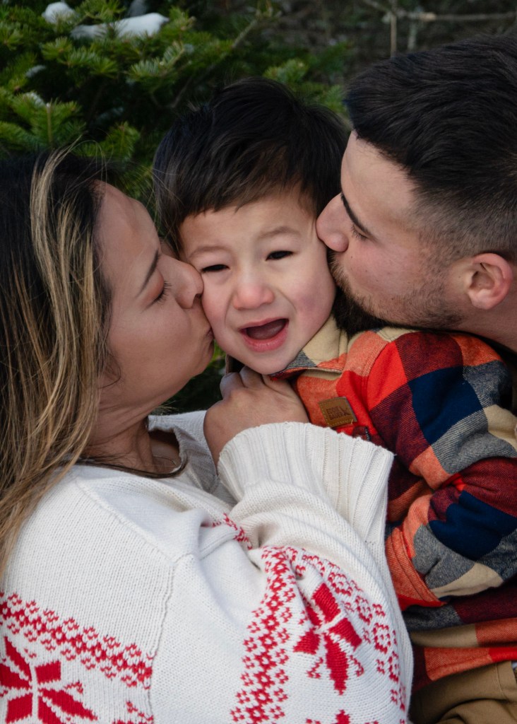 A family portrait featuring a woman and a man kissing a young child in between them. The child has a distressed expression while wearing a colorful plaid shirt. The adults are dressed warmly, with one wearing a white sweater adorned with red snowflake patterns, and the background shows greenery.