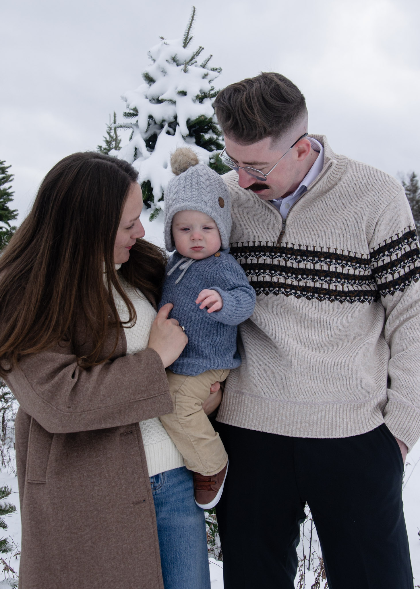 A family portrait featuring a woman and a man holding a baby, standing in a snowy landscape with evergreen trees in the background. The baby is wearing a gray hat and a blue sweater, while the adults are dressed in warm winter clothing.