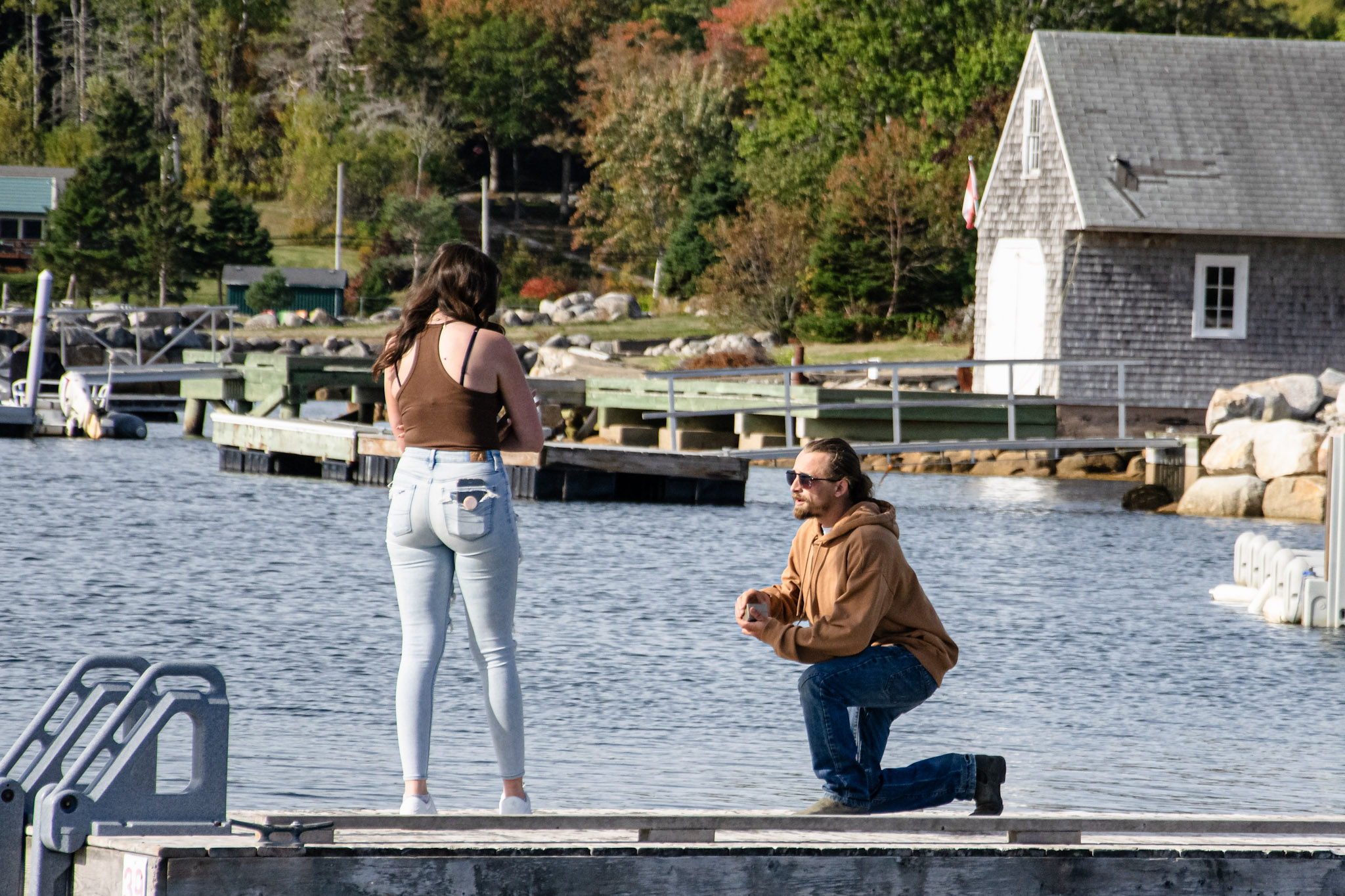 A man kneeling on a dock by a lake, proposing to a woman standing in front of him. The woman is wearing a brown tank top and light blue jeans, while the man is dressed in a brown hoodie and jeans. The background features trees with autumn-colored leaves and a small building.