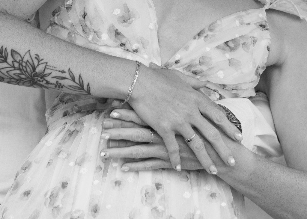 Close-up of intertwined hands on a floral dress, showcasing a tattoo on one arm and delicate rings on the fingers.