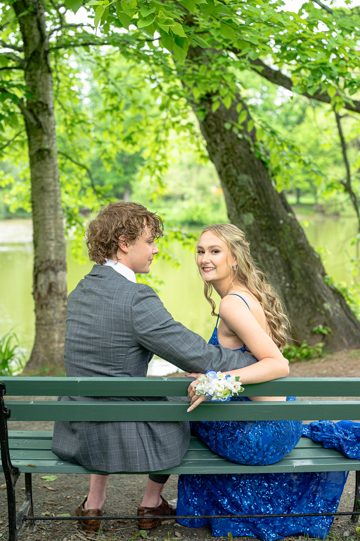 A couple sits on a park bench by a pond, smiling at each other. The woman wears a sparkling blue dress with a matching corsage, while the man is in a gray suit.