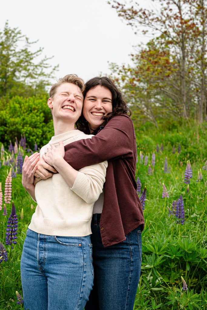 Two women smiling and embracing each other in a field of lupins, surrounded by greenery.