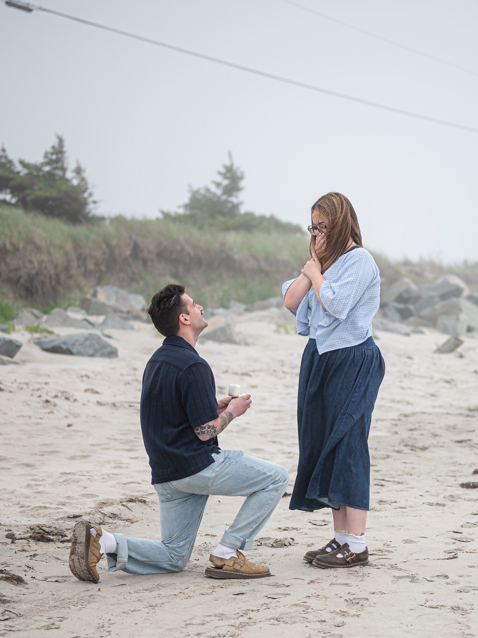 A man kneeling on a beach, proposing with an engagement ring to a surprised woman, who is covering her mouth in excitement.