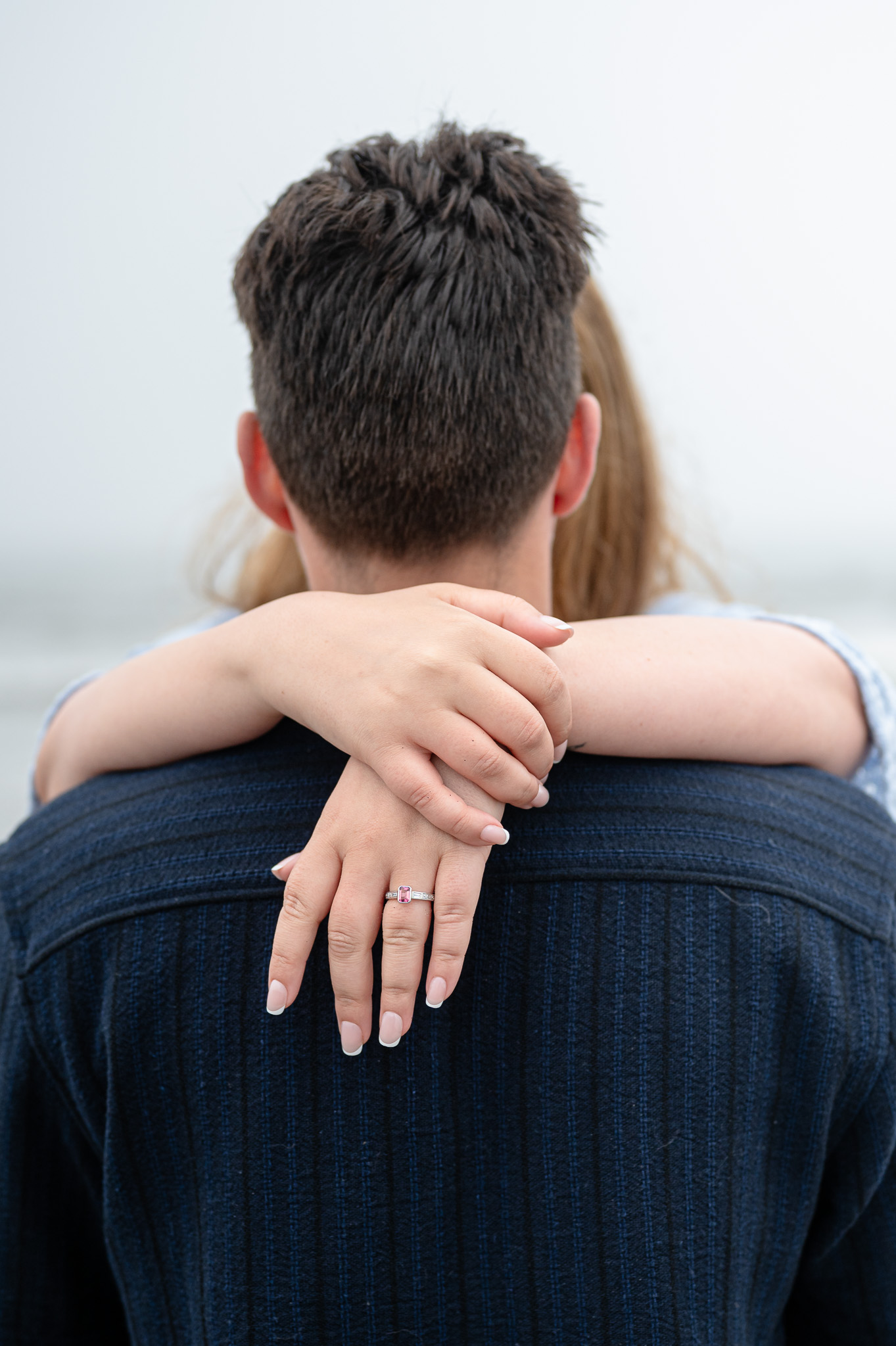 A couple embracing from behind, with a close-up on the woman's hand resting on the man's shoulder, showcasing an engagement ring.