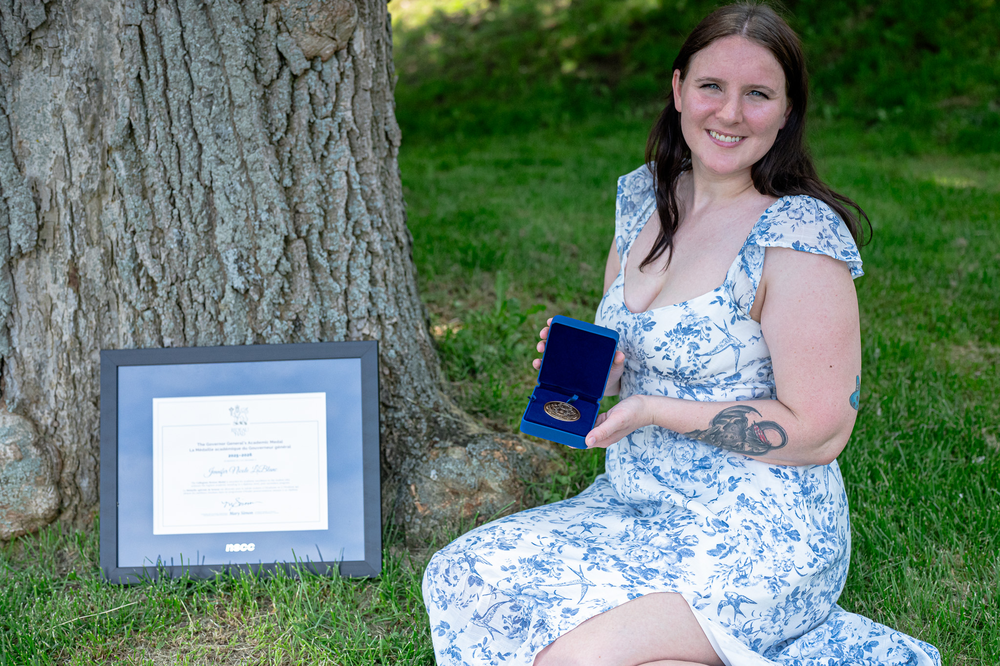 A woman sitting on the grass beside a tree, holding a medal in one hand and smiling. A framed certificate is positioned nearby on the ground.