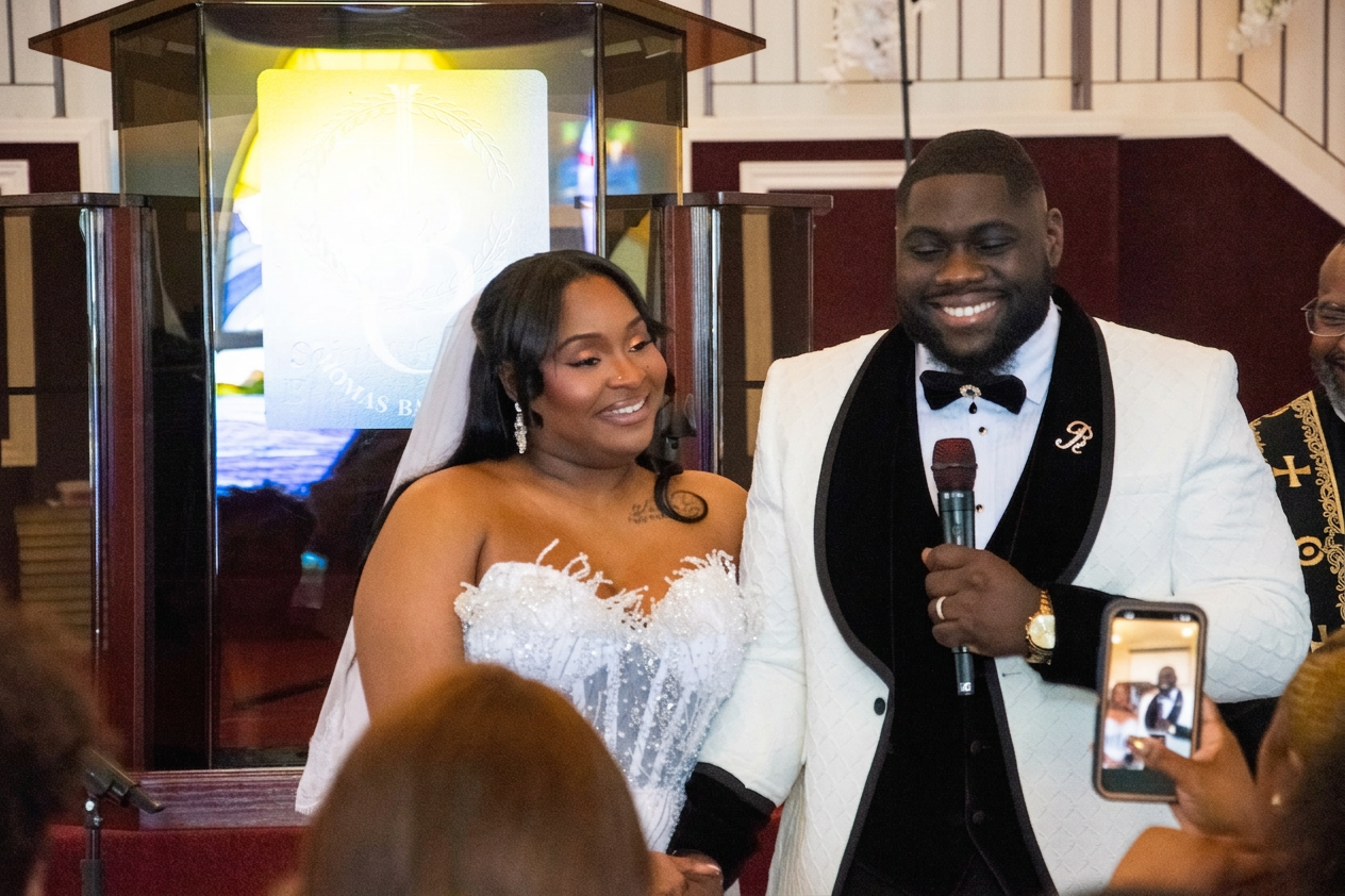 A couple stands together during their wedding ceremony, with the woman in a white gown and veil and the man in a formal black and white suit. They appear happy, sharing a moment as they address their guests, with a microphone in the man's hand.