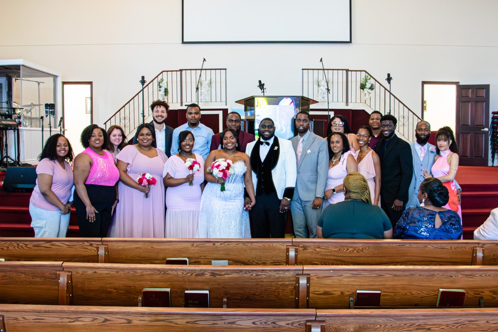 A wedding party poses together in a church. The bride, wearing a white dress, stands in the center holding flowers, surrounded by bridesmaids and groomsmen in various shades of pink and gray, all smiling.