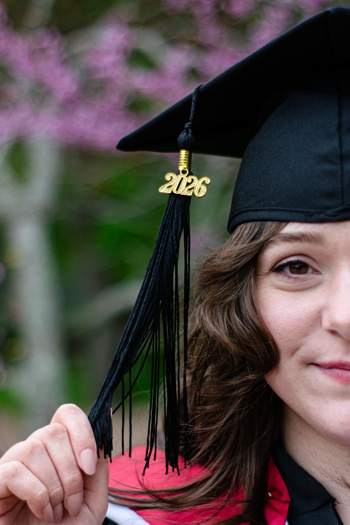 A close-up of a graduate wearing a black cap and gown, holding the tassel that features the year '2026'. The background is a blurred pink floral setting.