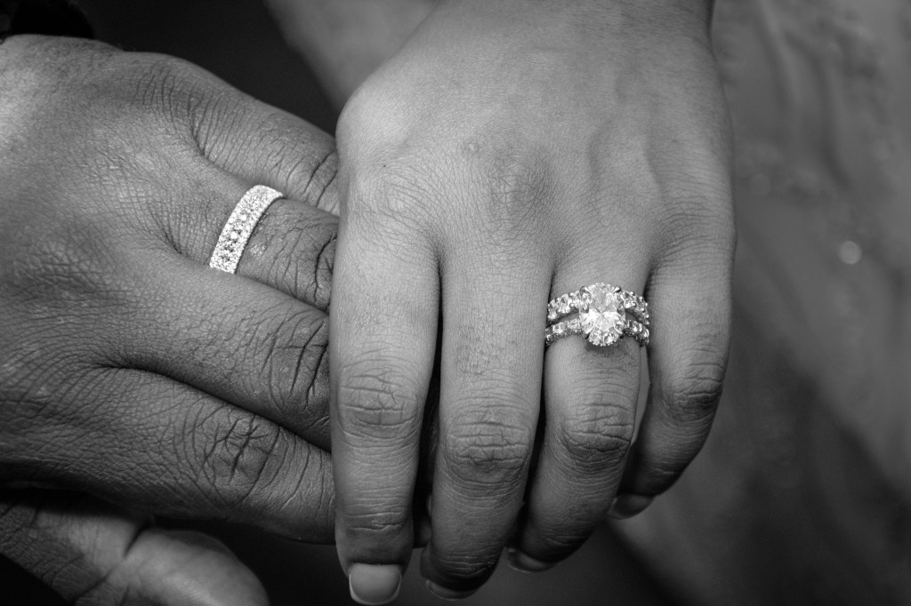 Close-up of two hands with wedding rings, one hand with a simple band and the other with an ornate engagement ring.