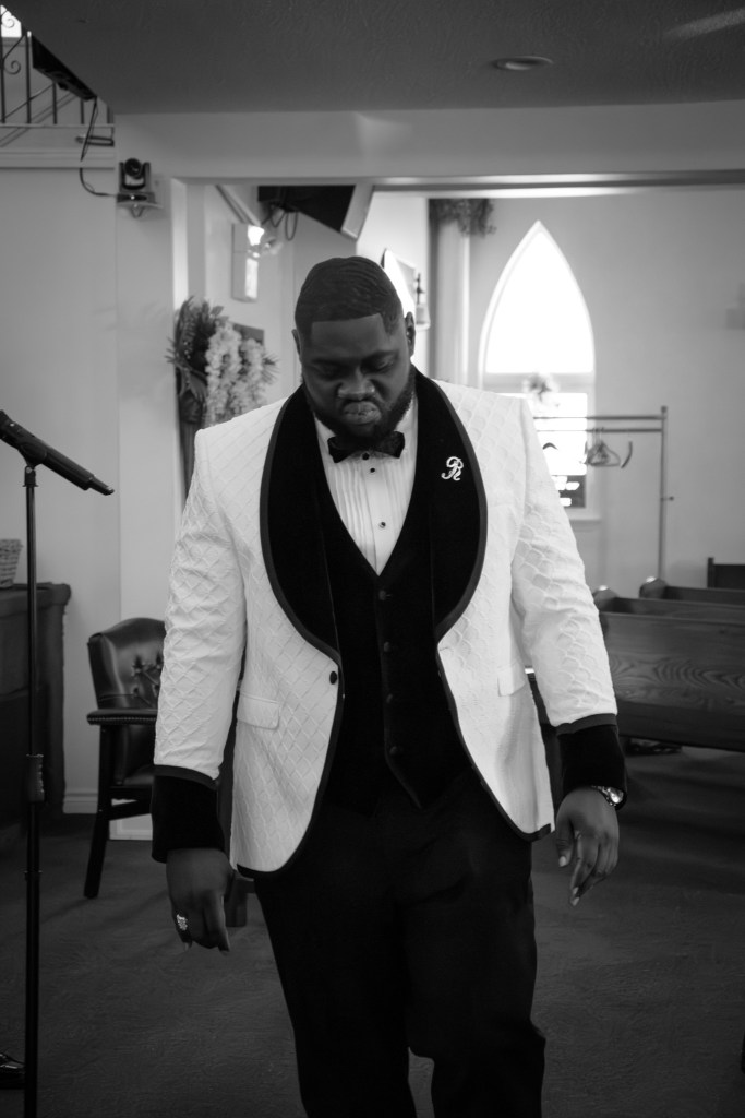 A man in a formal white tuxedo with a black bow tie walking in a church setting, focusing on the ground, captured in black and white.
