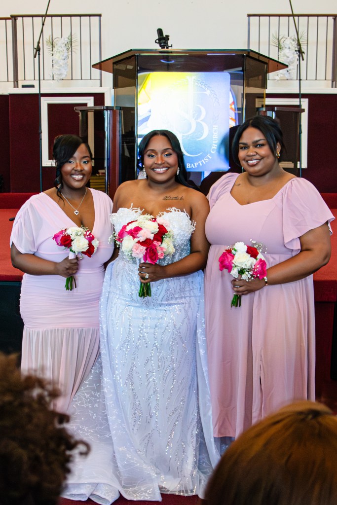 Bridal party posing together in a church setting, with the bride in a white gown and her friends in pink dresses, each holding bouquets of flowers.