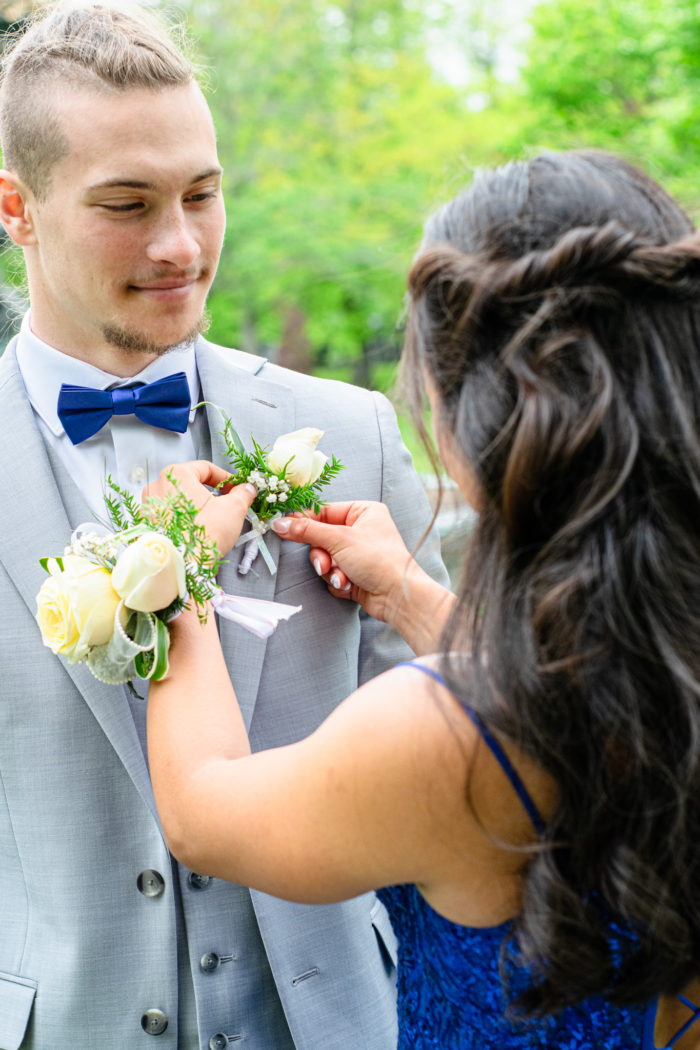 A woman pins a white rose boutonniere on a man's suit jacket during a formal event, surrounded by greenery.