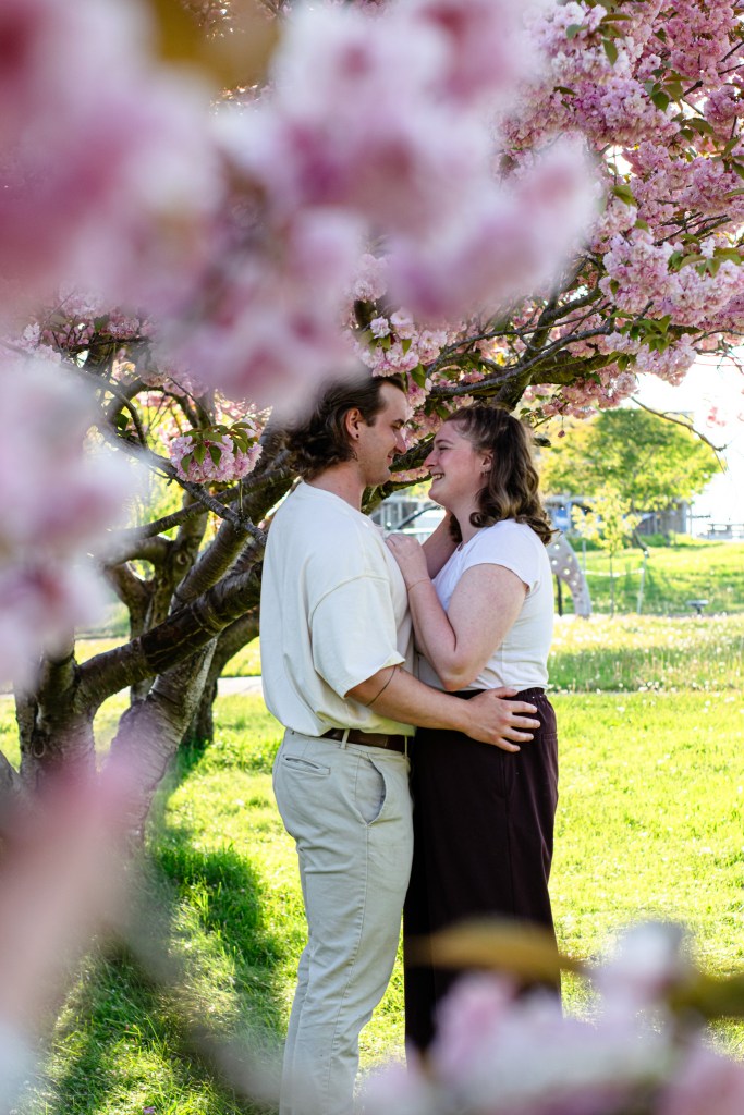 A couple embracing and smiling at each other under a cherry blossom tree, surrounded by pink flowers.