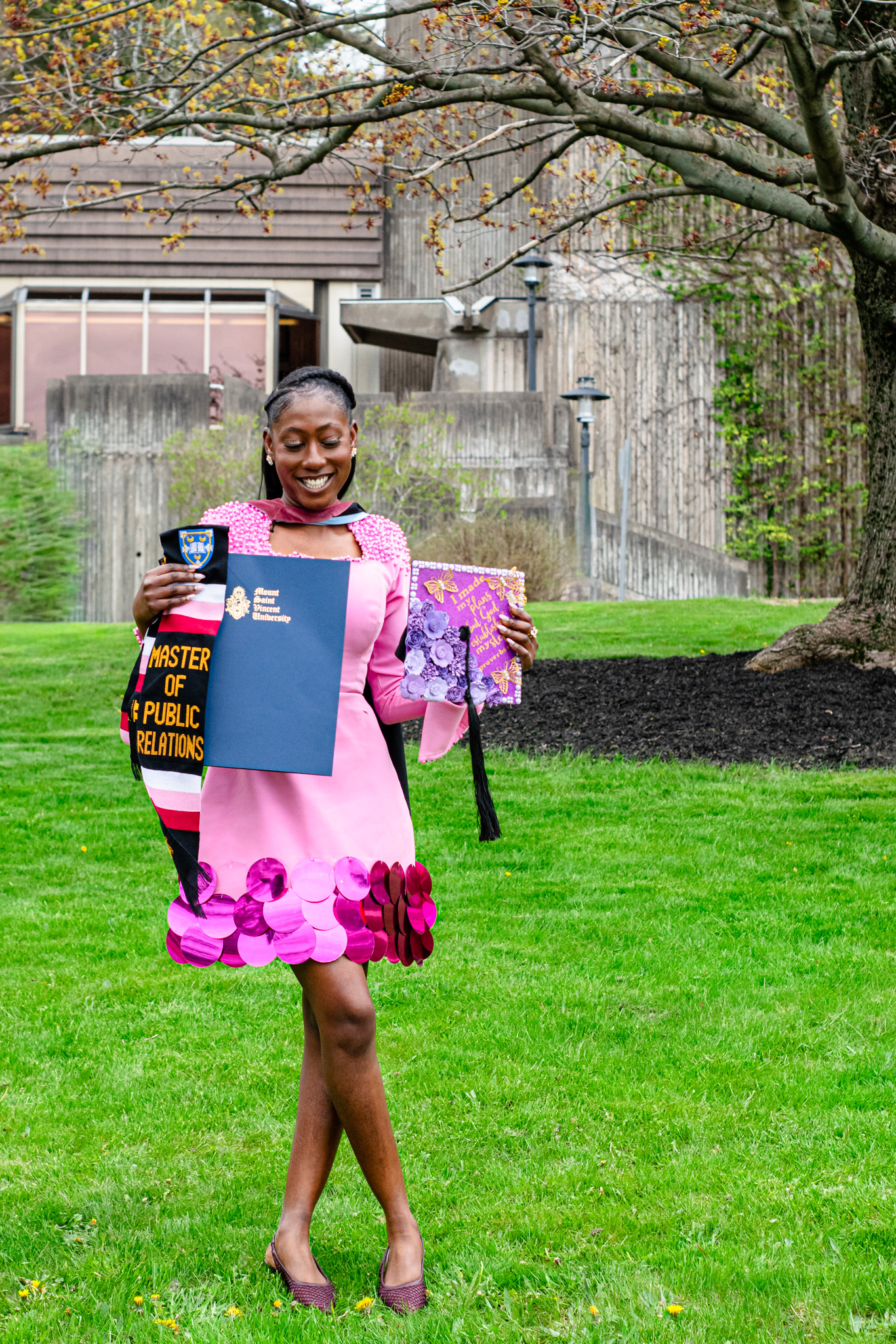 A woman in a pink dress with a decorative hem holds her diploma and graduation items in a green outdoor setting, smiling joyfully.