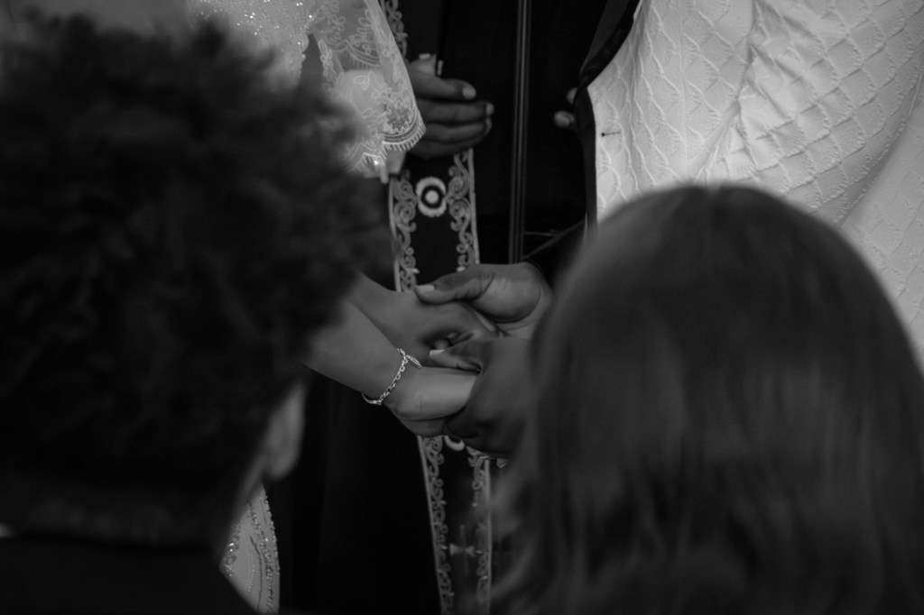 A black and white image of a wedding ceremony, focusing on two people holding hands during the exchange of vows, with guests' heads in the foreground.