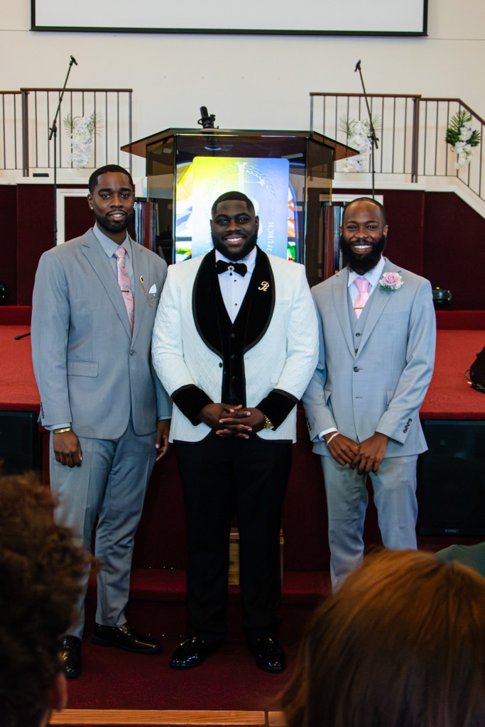 Three men posing together in formal attire inside a church, with a colorful backdrop and a podium in the background.