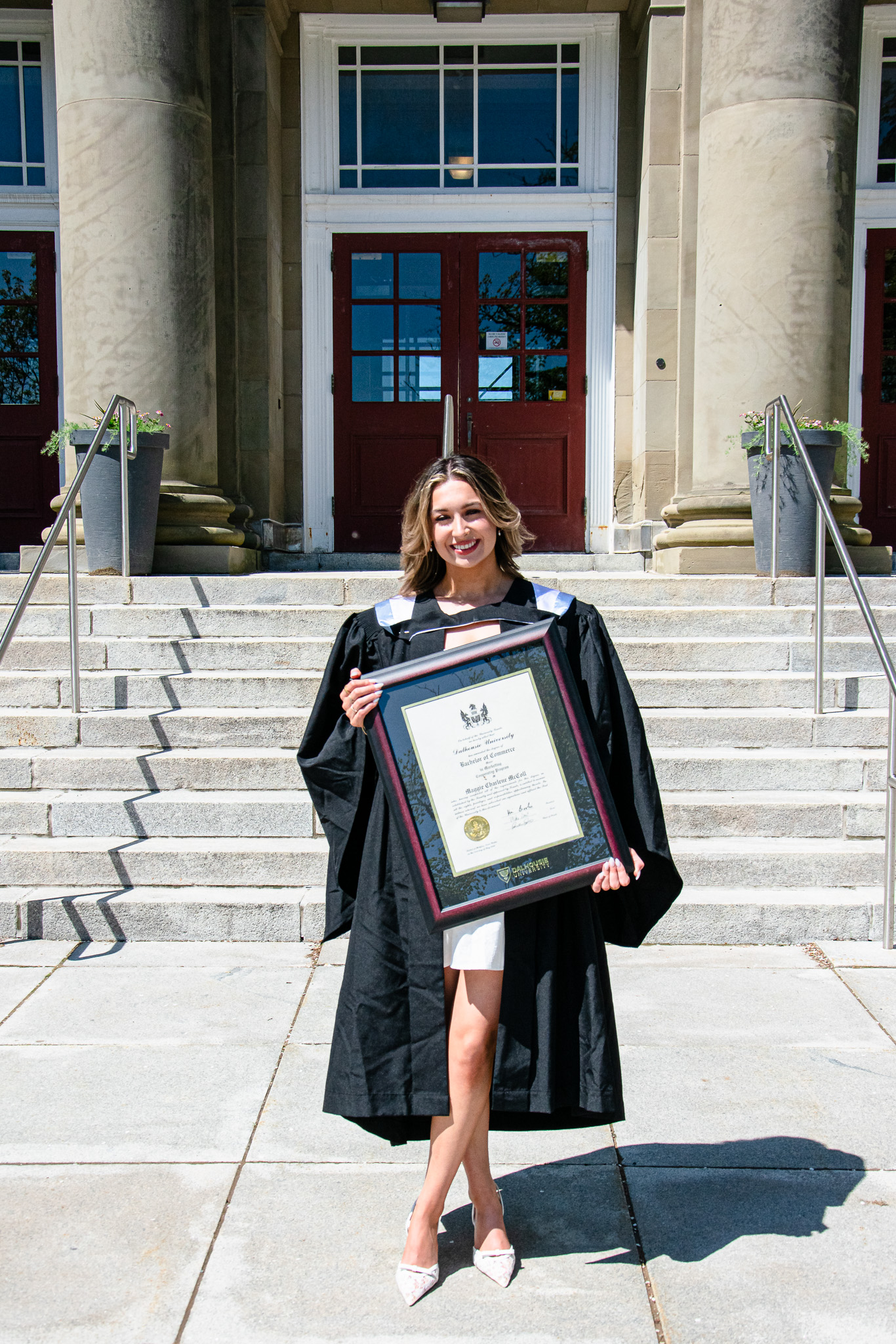A graduate in a black gown stands on the steps of a university building, holding a diploma framed in black. She smiles at the camera, showcasing her achievement.