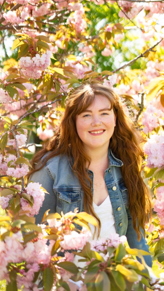 A woman with long, wavy hair smiles warmly while standing among blossoming pink cherry trees.