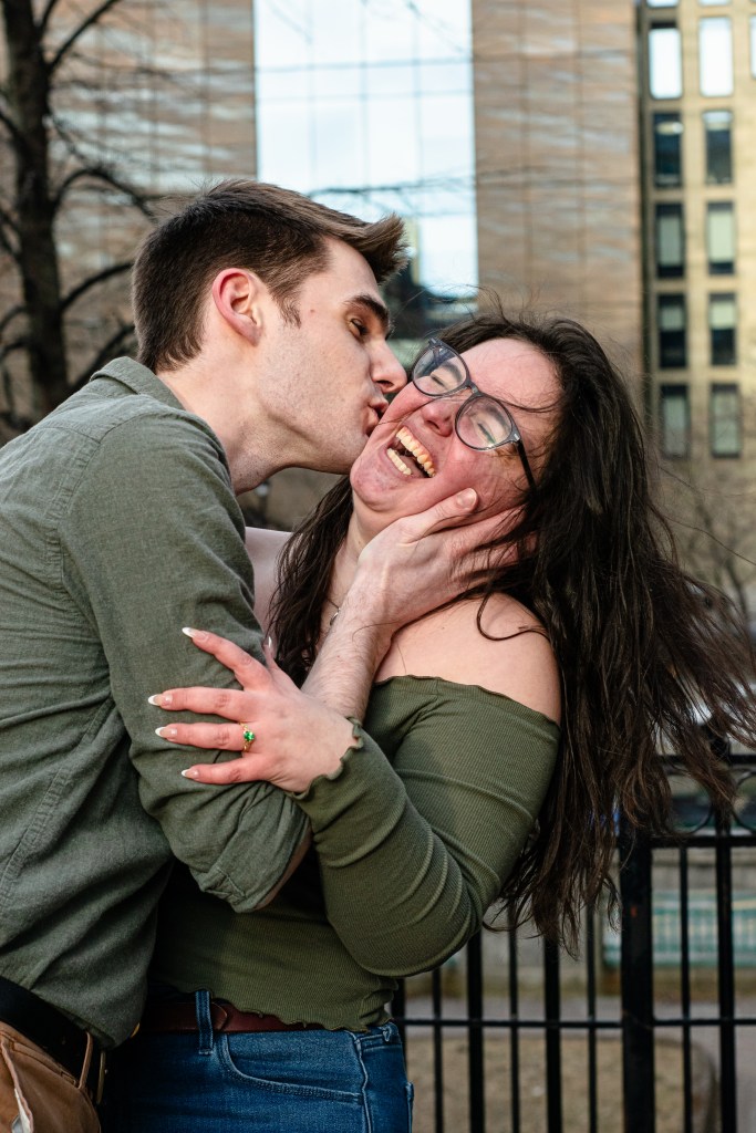 A man kisses a woman playfully while she smiles broadly, showcasing a joyful moment between them, with a blurred city backdrop.
