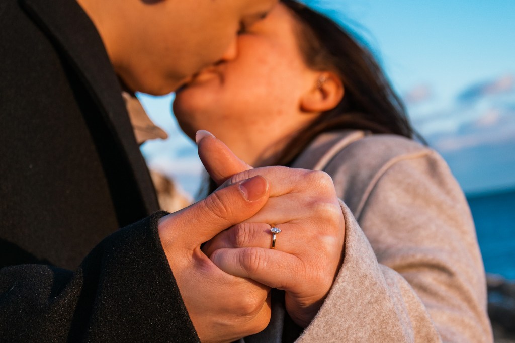 A romantic moment between a couple sharing a kiss, with a focus on their intertwined hands, showcasing an engagement ring against a scenic background.