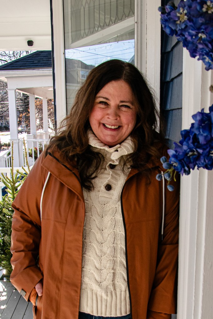 A woman smiling while standing on a porch, wearing a brown jacket and a cream-colored cable-knit sweater, with blue flowers in the foreground.