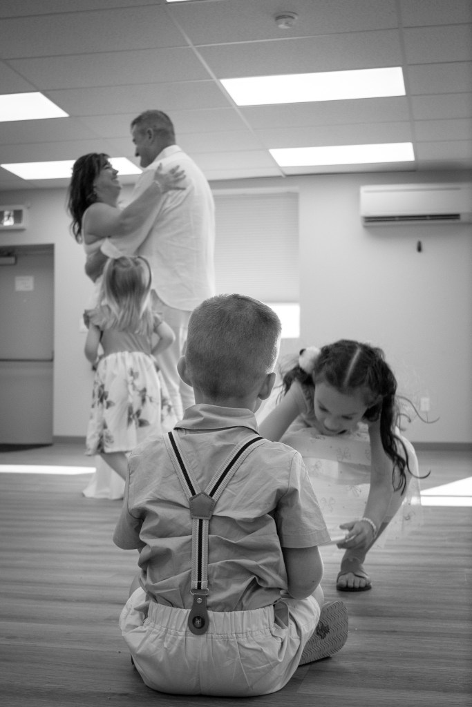 A black and white photo of a family gathering, with a couple dancing in the background while children play in the foreground. One boy in suspenders sits with his back to the camera, observing two girls nearby who are engaged in activities.