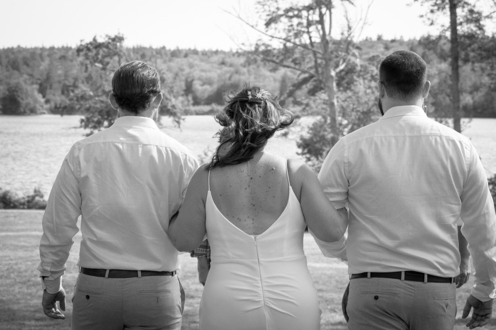 A bride in a white gown walking with two men, seen from behind, against a scenic outdoor backdrop of trees and water.