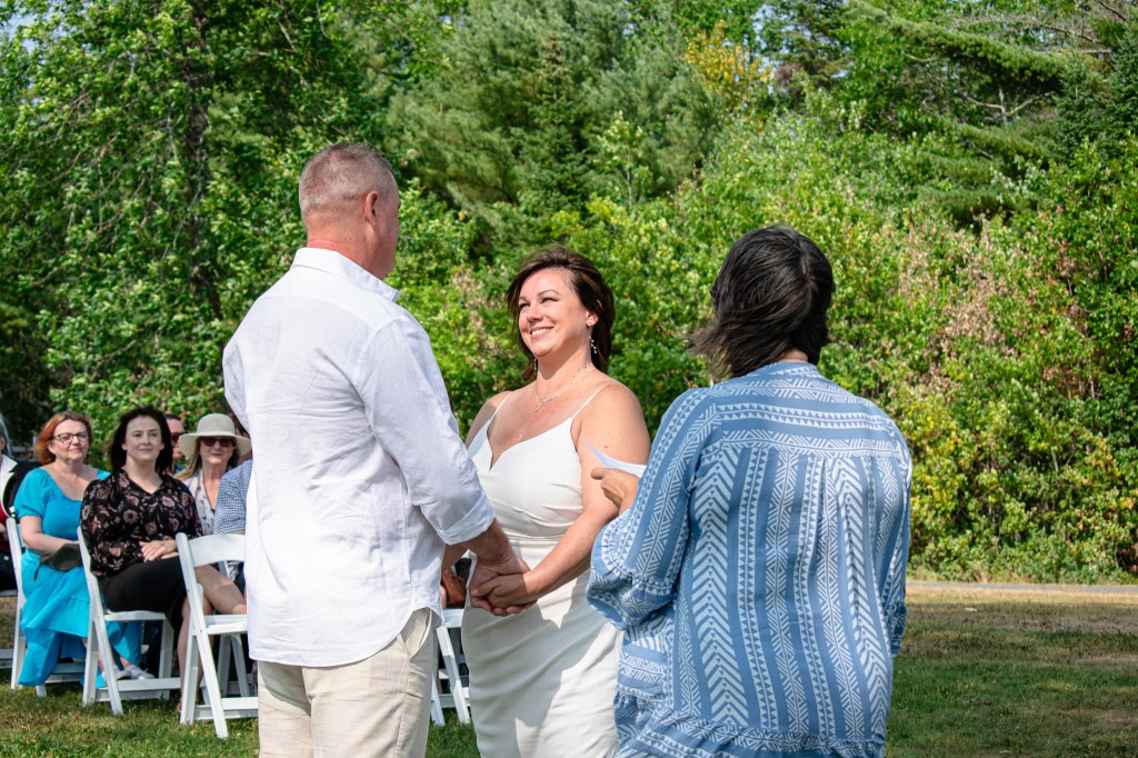A couple exchanges vows during an outdoor wedding ceremony, with guests seated in the background and greenery surrounding the scene.