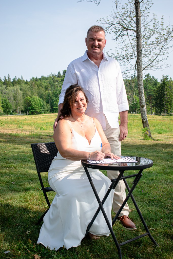 A couple standing together in a sunny outdoor setting, with the woman in a white dress seated at a table, signing documents, while the man in a light-colored shirt stands behind her.
