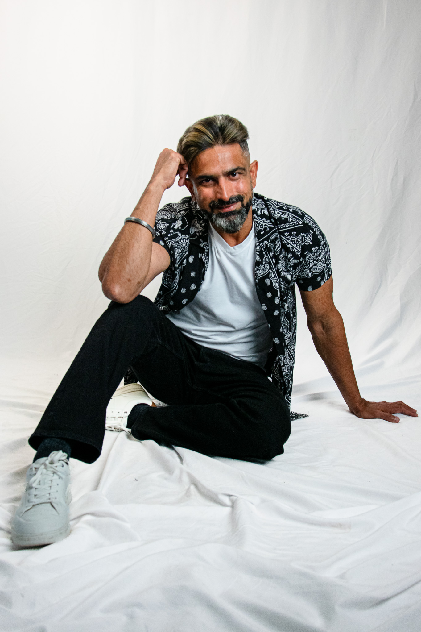 A man sitting on a white backdrop, wearing a patterned black shirt over a white t-shirt, black pants, and white sneakers. He is resting his chin on his hand with a slight smile.