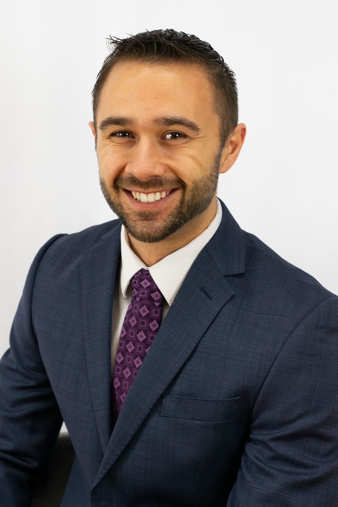 A professional man smiling, wearing a navy blue suit and a patterned purple tie, against a plain white background.