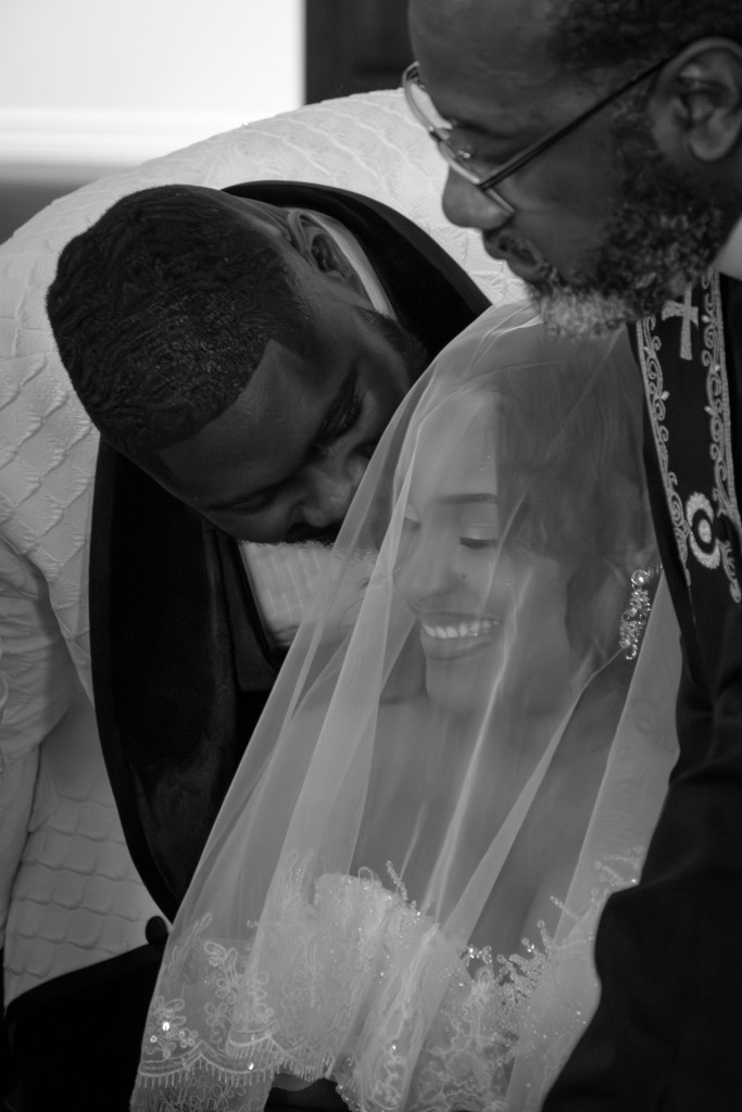 A close-up of a bride smiling under her veil as the groom lovingly leans in beside her, with a man in formal attire looking on, all captured in black and white.