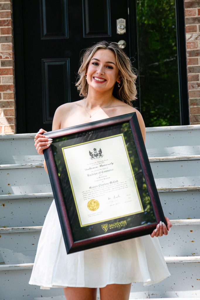 A young woman in a white dress stands on stairs holding a framed university diploma, smiling happily.