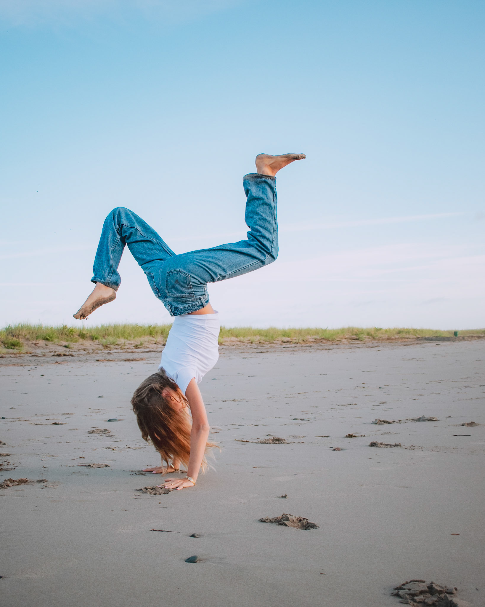 A young person performing a handstand on a sandy beach, wearing a white shirt and blue jeans, with a clear blue sky in the background.