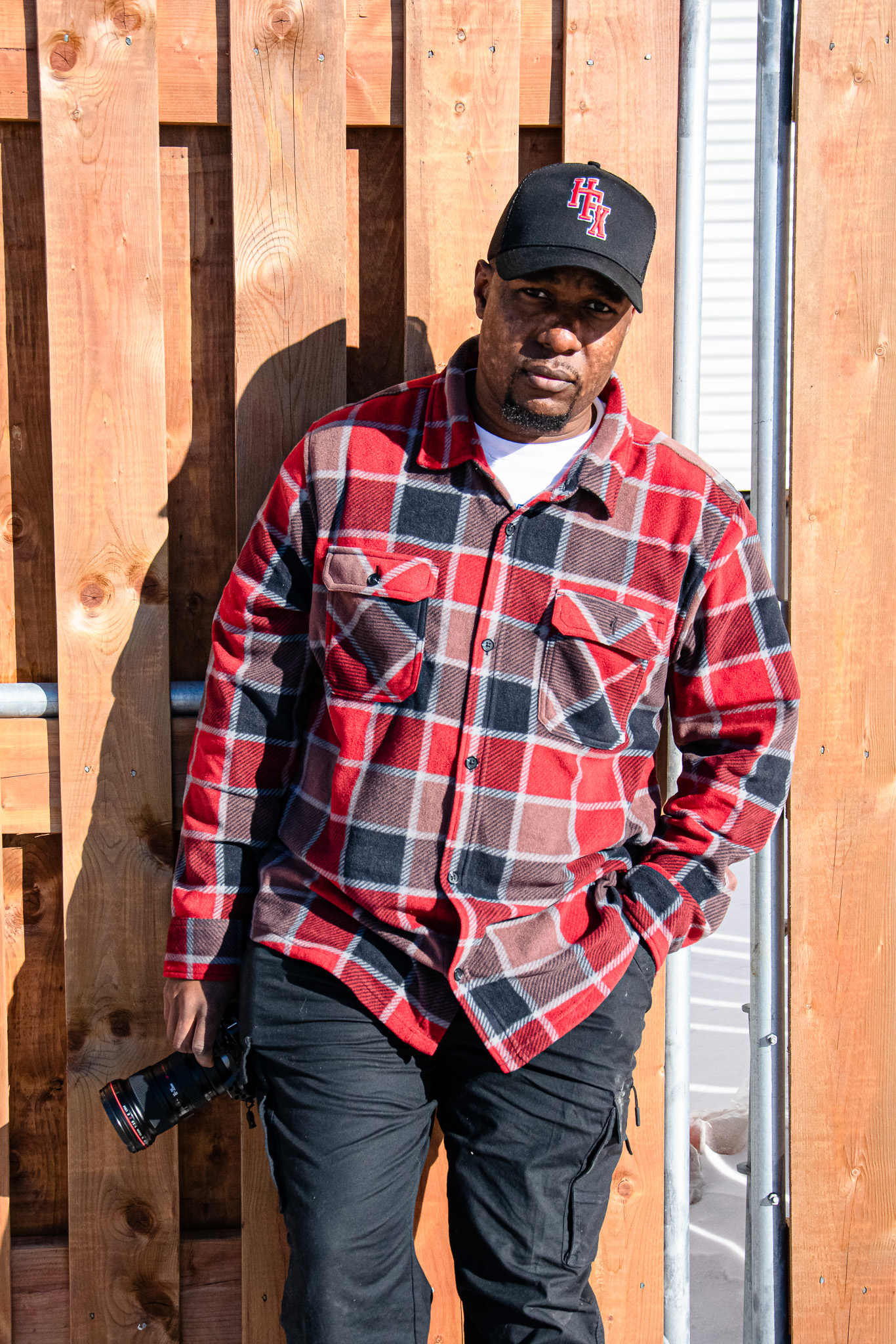 A man wearing a red and black plaid flannel shirt and a black cap stands against a wooden fence, holding a camera.