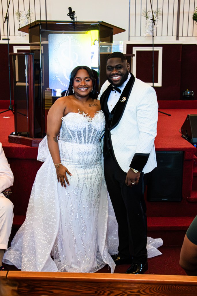 A smiling couple poses for a photo at a wedding ceremony. The woman wears an elegant, sparkling white gown, while the man is dressed in a stylish white tuxedo with a black bow tie.