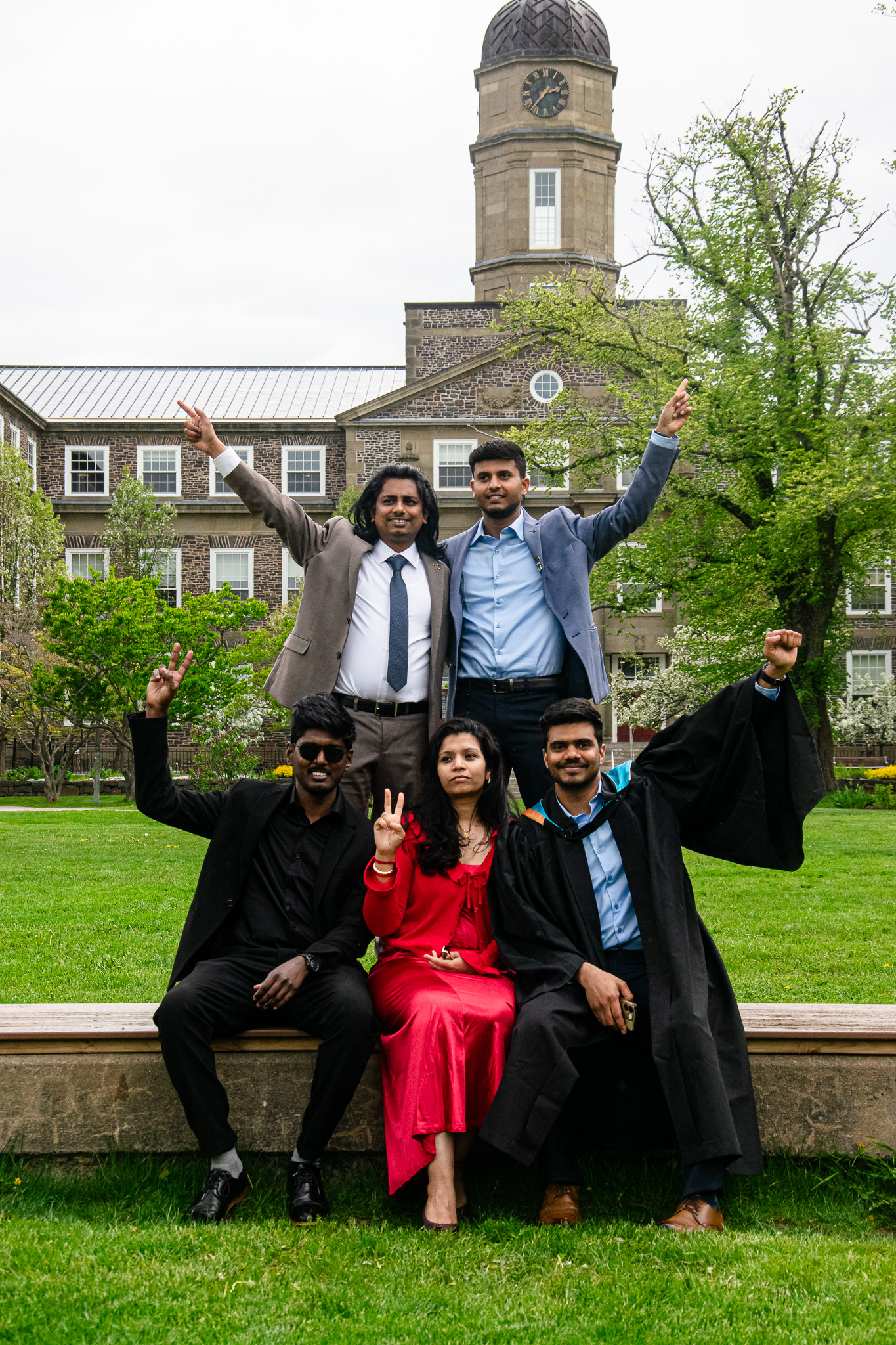 A group of five friends celebrating graduation outdoors, with two standing in the back making peace signs and three seated in front, one wearing a graduation gown. The background features a historic building and greenery.