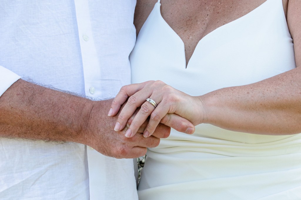 Close-up of two hands holding each other, one wearing a wedding ring, with a backdrop of light clothing.