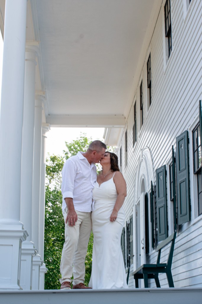 A couple sharing a kiss on the porch of a white wooden house, dressed in wedding attire.