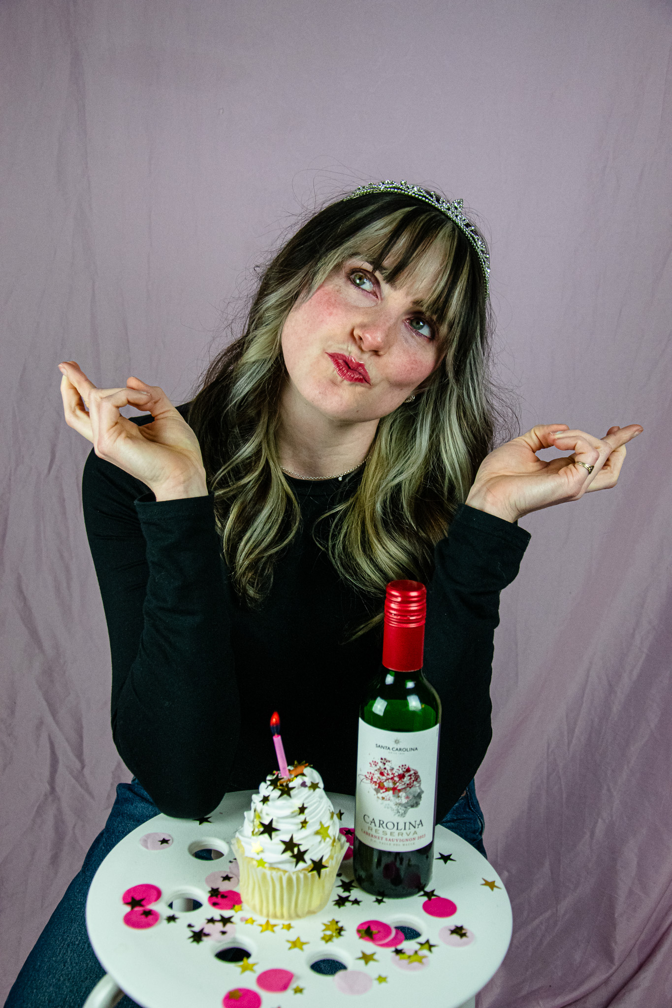 A woman wearing a tiara sits at a small table with a cupcake topped with a candle and a bottle of red wine, playfully making a funny face against a light pink background.