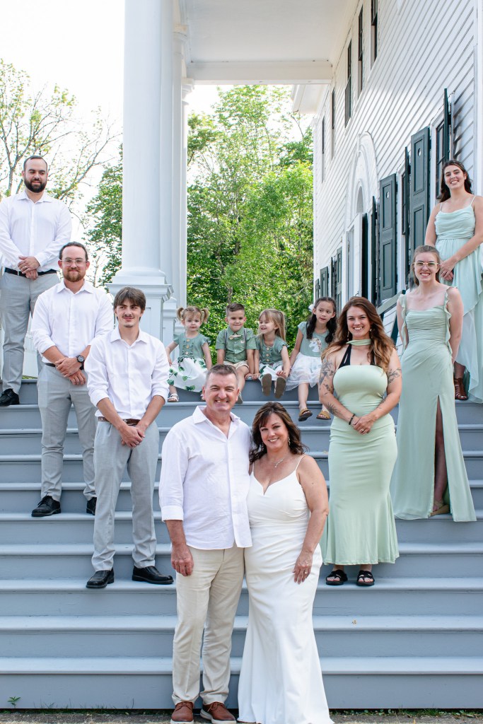 Wedding party posing on steps of a house, featuring a bride and groom in the foreground with family and friends arranged behind them.