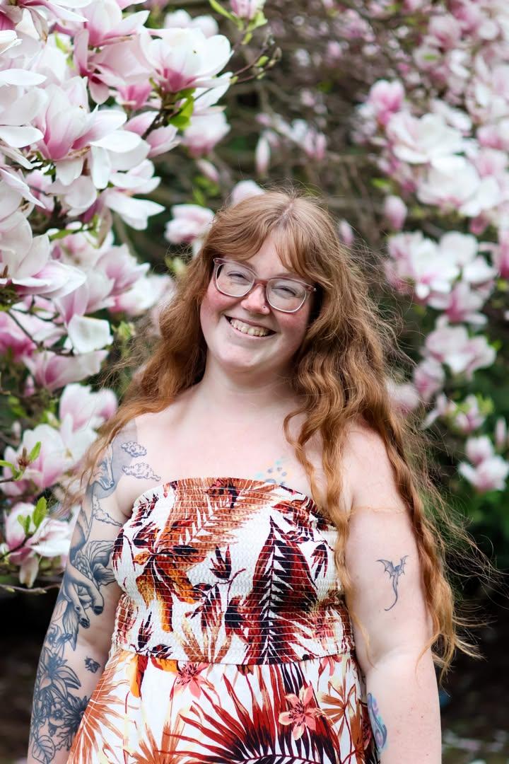 A smiling person with long curly hair, wearing a tropical-patterned dress, stands in front of blooming magnolia trees.