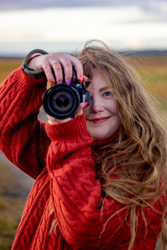 A woman with long curly hair wearing a red knitted sweater is holding a camera and smiling at the viewer.