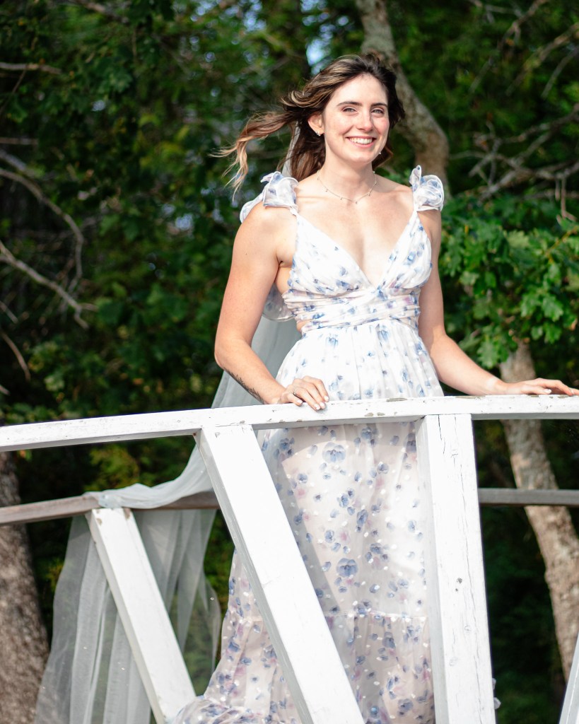 A smiling woman wearing a floral dress stands on a wooden structure surrounded by greenery.