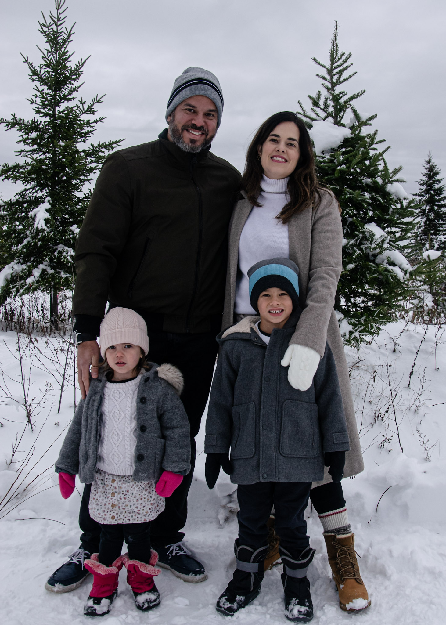 A family of four standing together in a snowy landscape, wearing winter clothing. The parents are smiling, with the father holding his daughter, while the son stands next to them. Snow-covered evergreen trees are in the background.