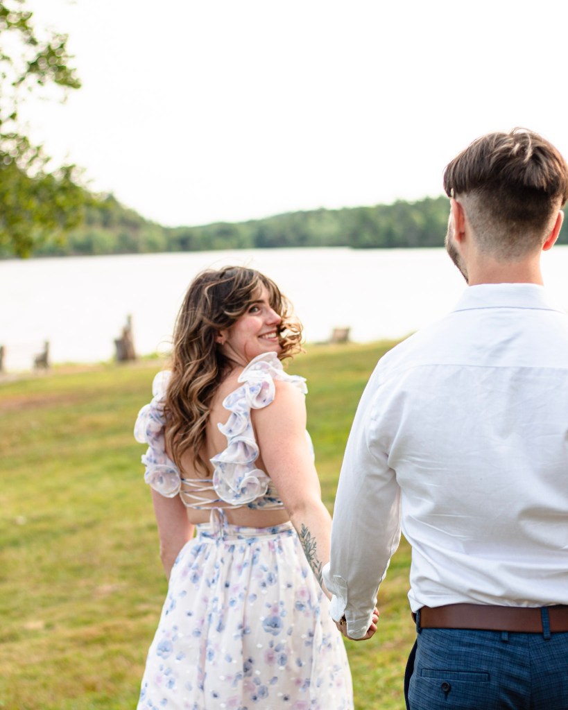 A woman with long, wavy hair smiles as she walks hand in hand with a man toward a lake, wearing a floral dress with ruffled sleeves.