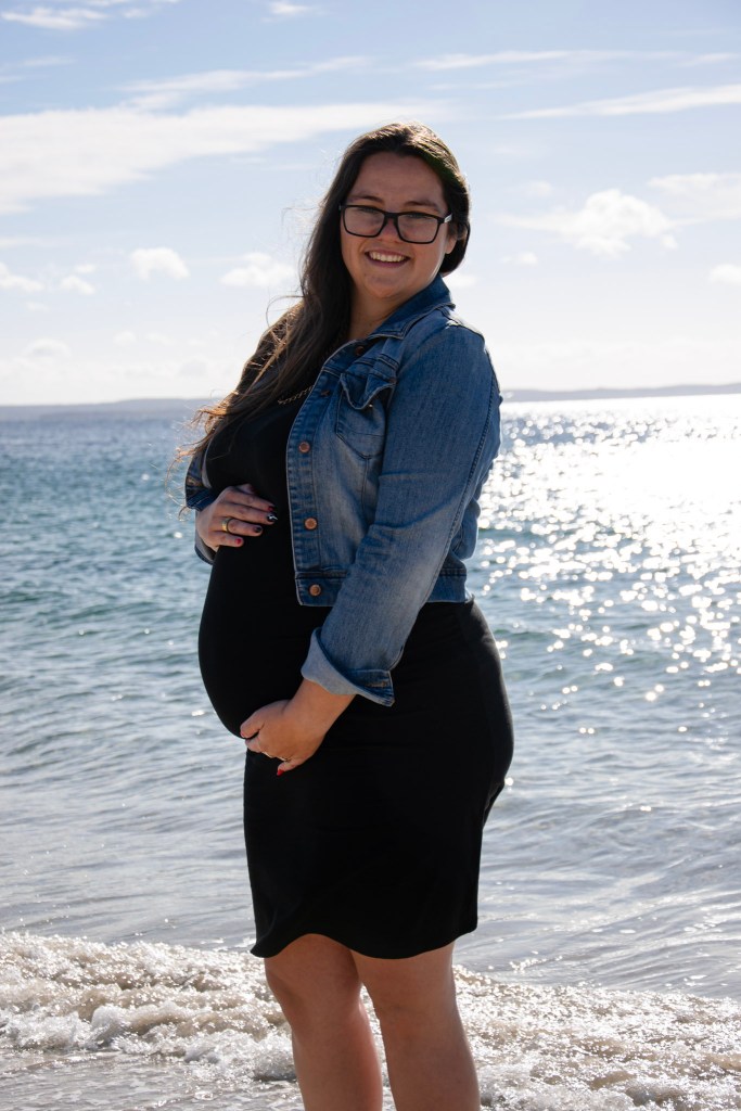 A pregnant woman standing on the beach, smiling at the camera, wearing a black dress and a denim jacket, with the ocean and a bright sky in the background.