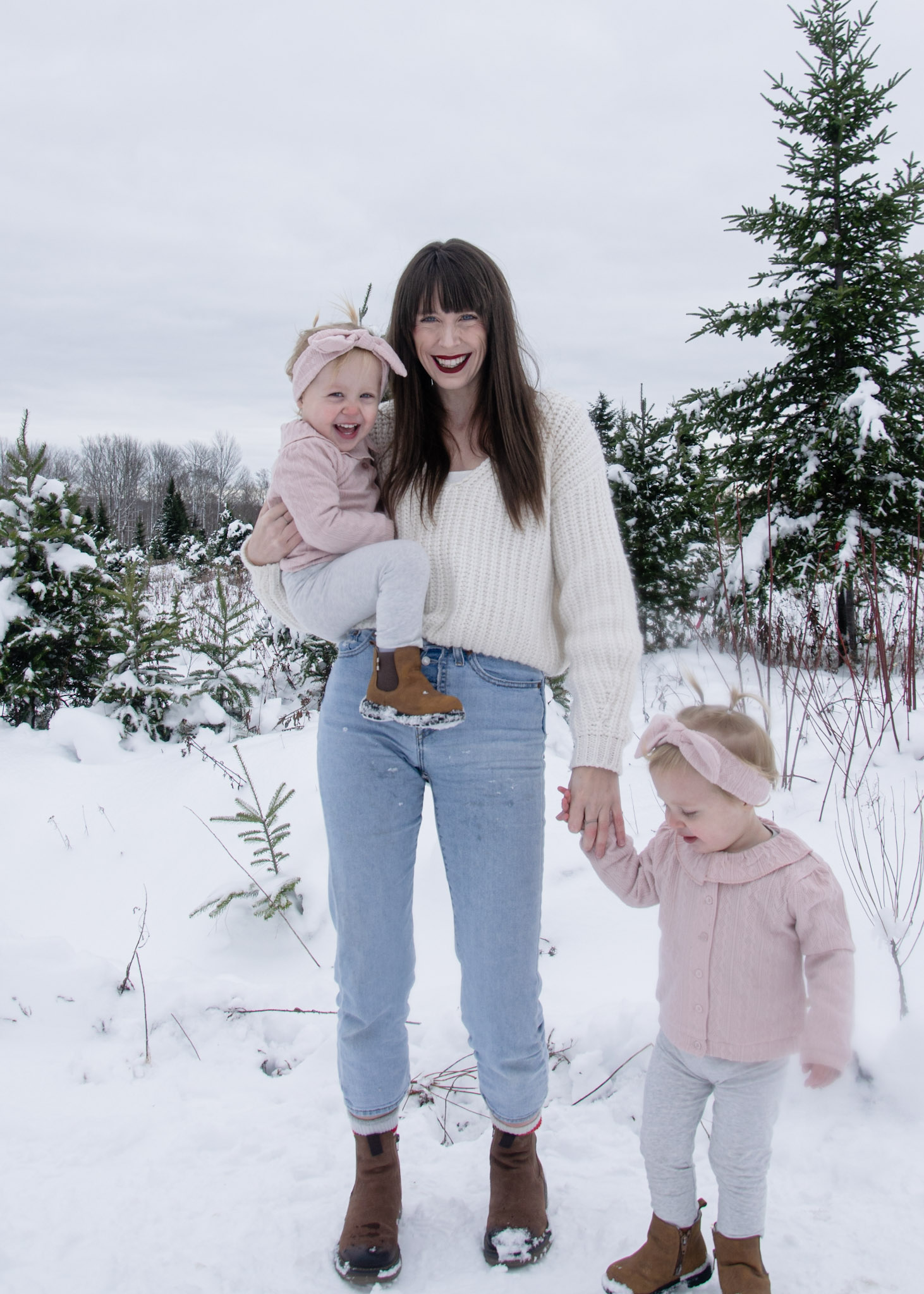 A woman wearing a cozy sweater and jeans holds a smiling toddler in a snowy landscape, while another young child walks beside her, holding her hand.